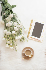 Notebook, tablet, a cup of coffee and a large bouquet white flowers on the floor on a white fur carpet. Freelance fashion comfortable femininity home workspace in flat lay style. 