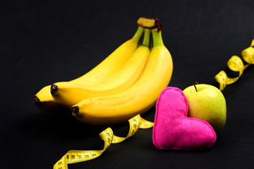 Bananas and apple near pink heart and tape measure