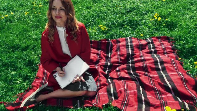Happy beautiful adult woman sits on the grass, writes in notebok, smiles. Outdoor, full body view