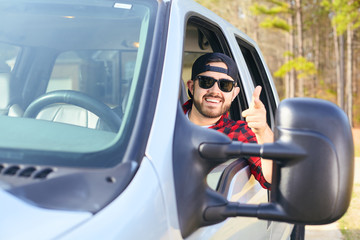 Handsome happy men driver with a beard smiling in the pickup car truck. Attractive male driving big vehicle, wearing hat, checkered shirt and black sun glasses. Sunny weather, summer