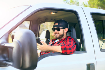 Handsome happy men driver with a beard smiling in the pickup car truck, looking at mobile phone, searching information in Internet, using social media. Attractive male driving big vehicle in hat.