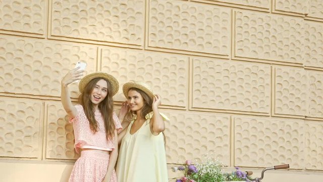 Young lovely friends women in hats and dresses making selfie on smartphone near bicycle in the street
