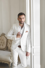 Fototapeta premium Elegant young handsome man with a beard in a white classic suit. The hotel's interior. Photo Studio. Sofa. Window. Chair. Chandelier
