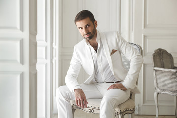 Elegant young handsome man with a beard in a white classic suit. The hotel's interior. Photo...