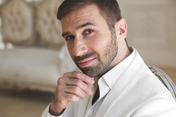 Elegant young handsome man with a beard in a white classic suit. The hotel's interior. Photo...