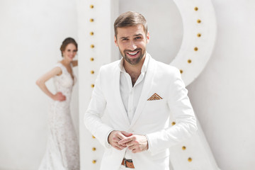 Fototapeta premium Chic wedding couple groom and bride posing in a white Studio. Happy couple laughing. White dress. White suit. Youth. Wedding. Chandelier. Sofa. The door.