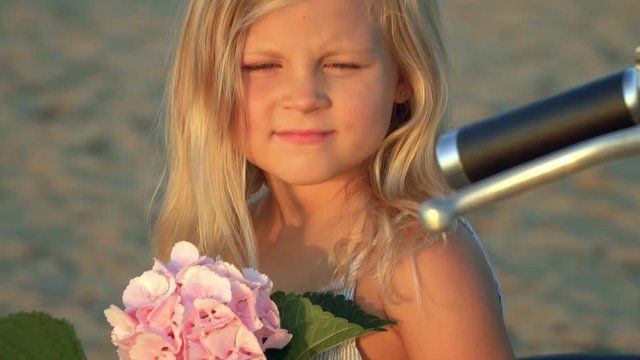 The little girl the blonde sits with a pink flower in hands on the white moped on sand by the sea. Child have amicably fun on vacation