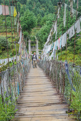 Fototapeta premium People walking on Bhutanese pedestrian suspension bridge with prayer flags in greenery area, Bhutan.