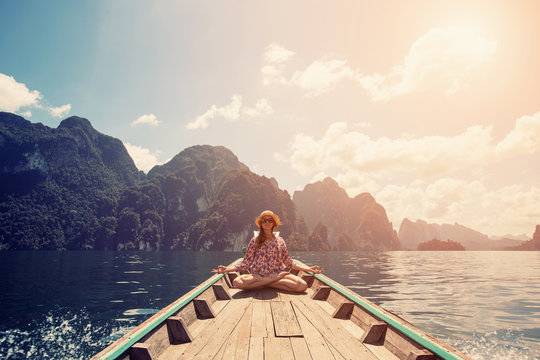 Young female sitting on the boat in yoga lotus pose and meditate. Travel on mountain lake, Thailand