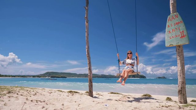 Young slim caucasian woman swings back and forth on a rope swing on white sand beach Tanjung Aan with turquoise water in Lombok, Indonesia. Shot with Sony a7s and Atomos Ninja Flame on sunny day.