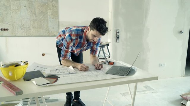 Young man working with smartphone and blueprints by table at his new home
