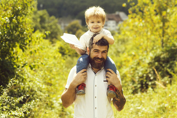Father, son plane in nature. Bearded parent holds on shoulders cheerful cute boy with paper airplane in hands. Walking and happy childhood, hobby aviation and family travel to airport. Chidhood dream