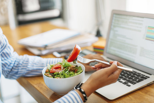 Cropped image of woman holding fork with tomato slice by laptop computer at wooden table