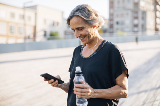 Happy woman looking at smart phone while holding water bottle