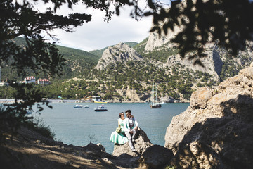 The bride and groom on nature in the mountains near the water. Suit and dress color Tiffany. Sitting on the stone.