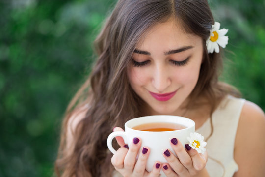 A beautiful young smiling woman with long curly hair in white top and a flower in hair holding a white cup of tea in hands, enjoying tea and looking down at the cup, green trees in the background