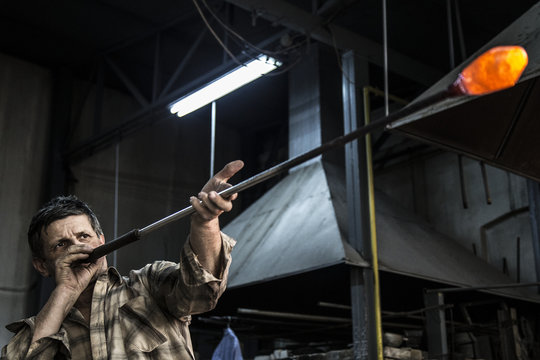 Man blowing molten glass with tube in factory