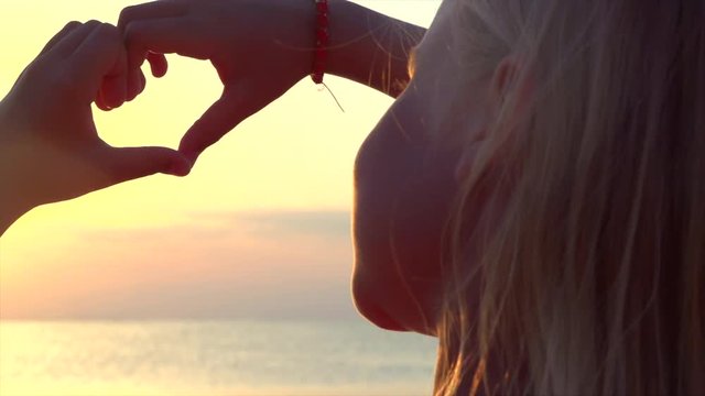 Happy little girl making heart with her hands over sunset sea background. Vacation concept. Summer holidays. 4K UHD video 3840x2160