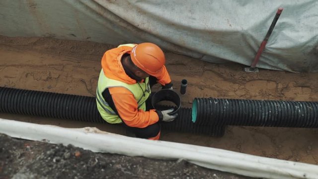 Worker in orange hard hat sitting in a ditch grease peace of black plastic tube