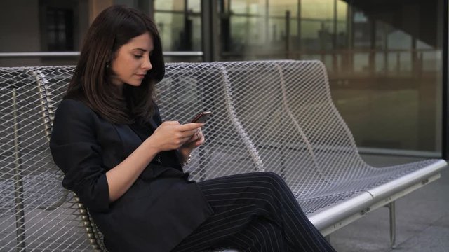 Side view of a young woman with long dark hair sitting on a rooftop in Madrid during a sunset. She is web sufring from her smartphone. Handheld real time medium shot