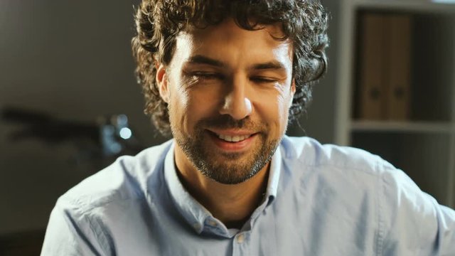 Portrait of handsome man with curly hair looking to the camera and smiling in the office background at night. Close up.