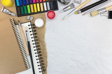 different drawing tools: sketchbook, watercolor, paintbrushes, colored chalks, gouache, pencils on recycled paper background