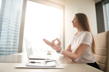 Calm peaceful businesswoman practicing yoga at office desk sitting near laptop, hand in chin mudra yogic gesture, meditation at work, online yoga exercises, practicing breathing, no stress, side view
