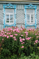 Fototapeta na wymiar Carved wooden platbands on the windows of the village house and flowers in front of the windows.