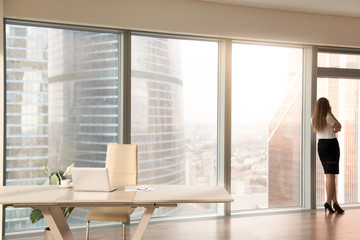 Modern office interior with desk and female silhouette standing back at full length window of business center, talking on phone, looking at cityscape, businesswoman having break at work after meeting