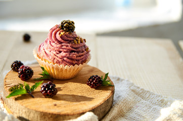 Homemade muffin in a brown baked basket with purple cream, fresh blackberry and golden splash decorated with raw blackberries and mint on wood frame on blurred gray background close up. Side view