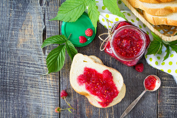 Homemade jam. Flat lay, top view. Glass jar with red raspberries and white bread on the kitchen table. Preserved berry.