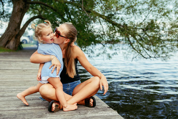 Fototapeta na wymiar Group portrait of happy white Caucasian mother and daughter child having fun outside. Mom and child girl playing, hugging kissing in park on pier by water lake. Candid authentic real lifestyle.