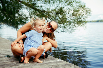 Fototapeta na wymiar Group portrait of happy white Caucasian mother and daughter child having fun outside. Mom and child girl playing, hugging in park on pier by water lake. Candid authentic real lifestyle.