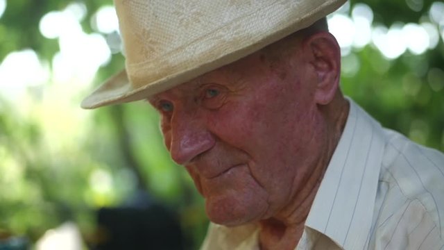 Very old man portrait with emotions. Grandfather in white hat sad and depressed. Portrait: aged, elderly, loneliness, senior. Close-up of a pensive old man sitting alone outdoors. Slow motion.