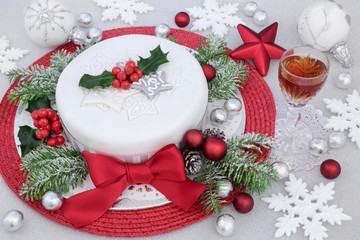 Iced Christmas cake with red bow, sherry drink, bauble and snowflake decorations, winter greenery and silver foil wrapped chocolates on glitter background.