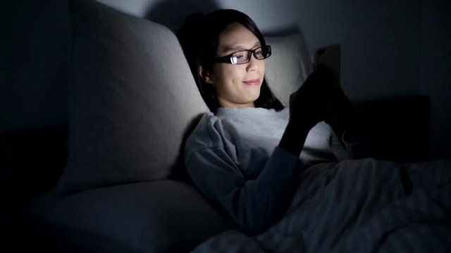 Woman wearing glasses and using cellphone on bed at night