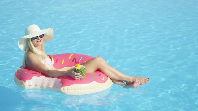 SLOW MOTION CLOSE UP Happy Blonde Girl Sipping Pineapple Drink And Relaxing On Inflatable Pink Doughnut Float. Young Woman In Bikini Enjoying Summer Vacation Drinking Cocktails On Pink Floatie In Pool