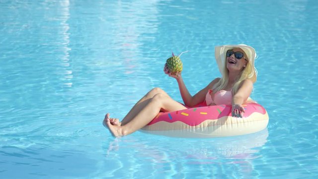 SLOW MOTION CLOSE UP Happy blonde girl sipping pineapple drink and relaxing on inflatable pink doughnut float. Young woman in bikini enjoying summer vacation drinking cocktails on pink floatie in pool