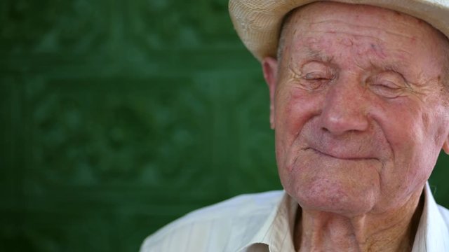 Very old man portrait with emotions. Grandfather happy and smiling. Portrait: aged, elderly senior. Close-up of a pensive old man in white hat sitting alone outdoors at summer. Slow motion.