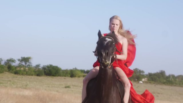 Young girl in long red dress riding black horse in countryside. Female rider with her stallion galloping across a field kicking up dust. Horseback riding in slow motion.