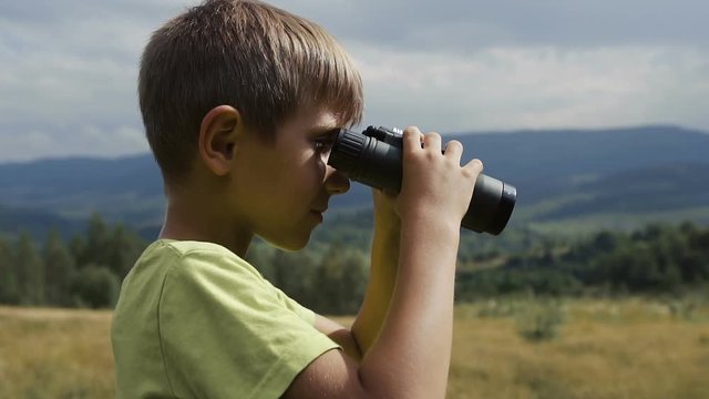 The boy stands on the top of the mountain and looks at the binoculars