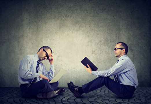 Two men sitting on a floor one reading a book another working on laptop