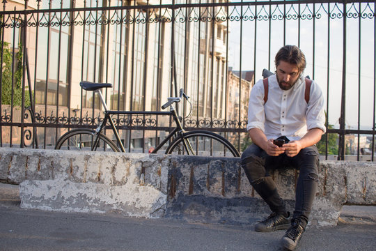 Young handsome mrn with beard sitting with cellphone on a bridge with bicycle on background  