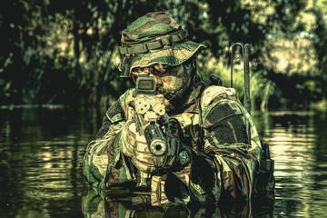 Bearded soldier in action during river raid in the jungle pointing rifle to camera. He is waist...