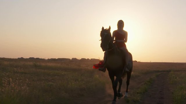 Young girl in long red dress riding black horse against the sun. Beautiful female rider with her stallion trotting across a field at sunset. Horseback riding in slow motion.