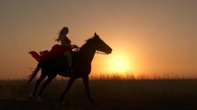 Young girl in red dress riding black horse against the sun. Rider with her stallion trotting across a field at sunset. Long gown blowing in the wind. Horseback riding in slow motion.