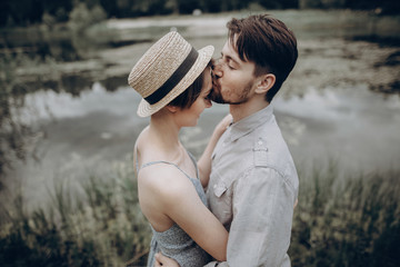 stylish hipster couple kissing at lake. man and woman embracing, in love relaxing in summer park, picnic date. girl in fashionable modern dress and straw hat . rustic wedding concept