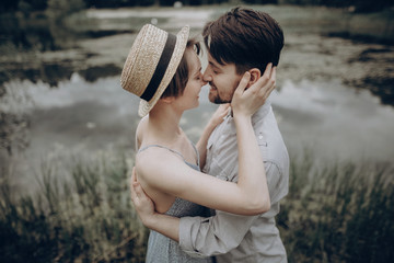 Fototapeta premium stylish hipster couple kissing at lake. man and woman embracing, in love relaxing in summer park, picnic date. girl in fashionable modern dress and straw hat . rustic wedding concept