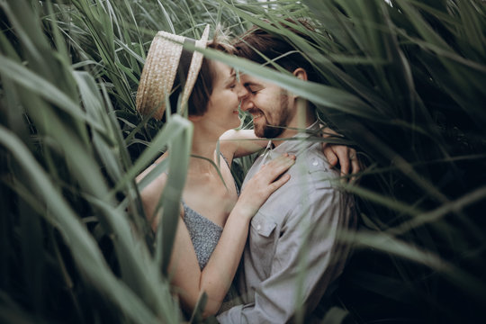 happy hipster couple embracing at lake in cane. stylish rustic bride and groom, girl in fashionable modern dress and straw hat smiling at river in windy high reed. rustic wedding concept.
