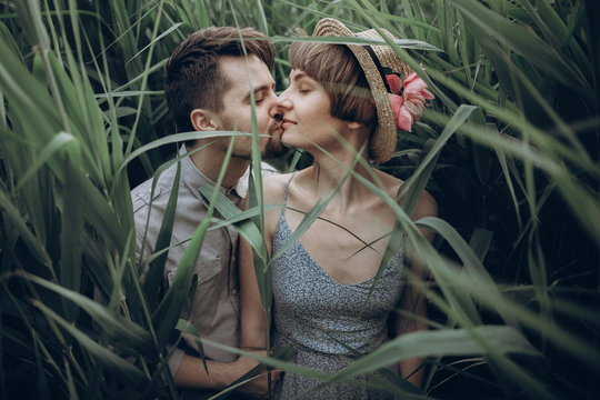 hipster couple posing at lake in cane in summer forest. stylish rustic bride and groom, girl in fashionable modern dress and straw hat with peony, embracing in windy high reed. rustic wedding concept